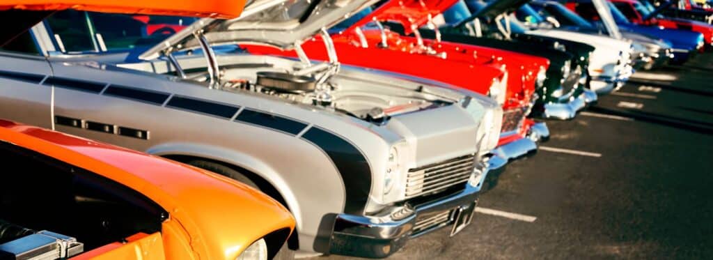 A selection of classic cars parked in line together displaying their engines via open bonnets. 