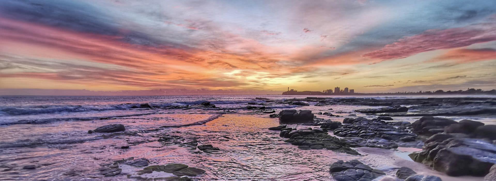 A dramatic seascape at sunset with a rocky coastline in the foreground. Dark volcanic or weathered rocks are scattered across the beach and extend into the churning ocean waters. The sky is filled with sweeping clouds painted in vibrant colors - deep blues and purples at the top transitioning to brilliant oranges, pinks, and yellows near the horizon. The wet sand and tide pools between the rocks reflect the colorful sky. In the distance on the right, a city skyline with tall buildings is silhouetted against the sunset. Waves crash against the rocks, creating white foam and spray.