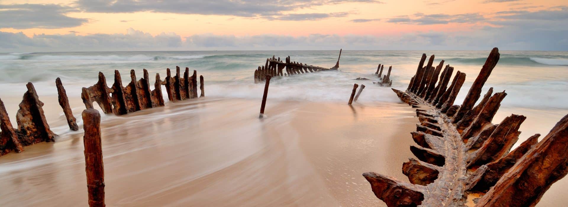 The skeletal remains of a shipwreck emerge from a sandy beach at sunset. Weathered wooden ribs and metal framework of what appears to be an old vessel create dramatic silhouettes against the moody sky. The long exposure photography captures the smooth, milky motion of waves washing around and through the wreck structure. Rusted metal posts and deteriorating wooden beams stick up from the sand at various angles, showing decades of decay from salt water and weather. The sky is filled with soft clouds in muted tones of gray, peach, and cream, creating an atmospheric and haunting coastal scene.