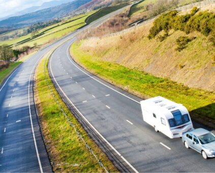 Aerial view of a white car towing a white caravan traveling on a curved highway through rolling countryside. The road winds through green hills and fields with autumn-colored vegetation, scattered trees, and mountains visible in the distance under a partly cloudy sky.