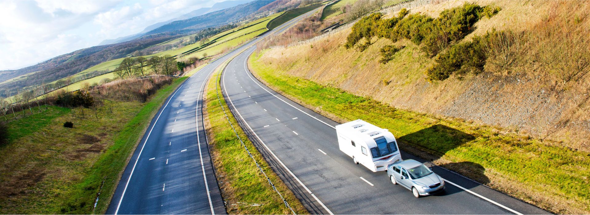 Aerial view of a white car towing a white caravan traveling on a curved highway through rolling countryside. The road winds through green hills and fields with autumn-colored vegetation, scattered trees, and mountains visible in the distance under a partly cloudy sky.