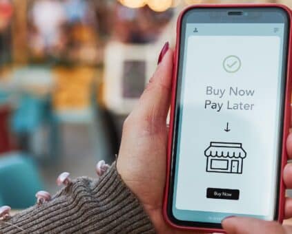 A hand holding a smartphone displaying a "Buy Now Pay Later" app interface with a checkmark, shop icon, and "Buy Now" button, set against a blurred background of colorful cafe or restaurant seating.
