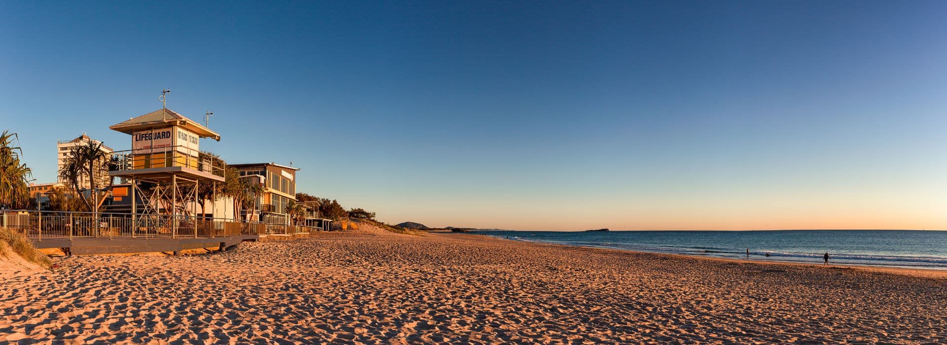 A golden hour beach scene showing a wooden lifeguard tower on stilts with "LIFEGUARD" signage, positioned on sandy beach with gentle waves. The beach stretches into the distance where a few people can be seen walking. Behind the lifeguard station are modern beachside buildings and palm trees. The sky transitions from deep blue at the top to warm golden tones near the horizon, creating beautiful lighting across the sand and water.