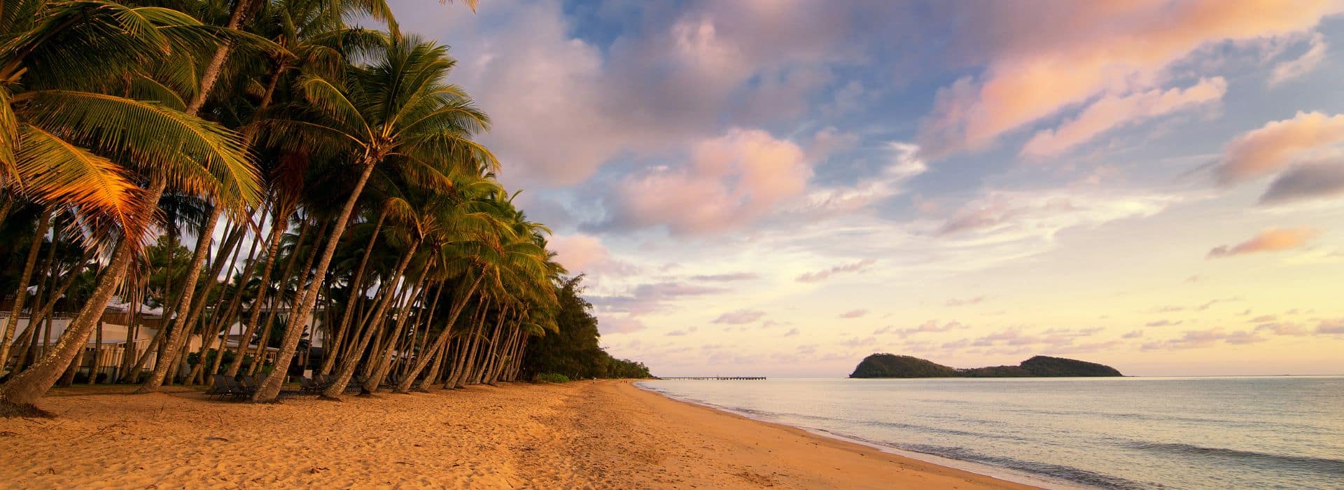 A tropical beach at sunset with a row of tall coconut palm trees leaning over golden sand. The palm fronds are illuminated by warm orange and pink light from the setting sun. The calm ocean stretches to the horizon where two small forested islands are visible in the distance. The sky is filled with soft clouds painted in pastel hues of pink, purple, and orange. The beach appears pristine and empty, with gentle waves lapping at the shoreline.