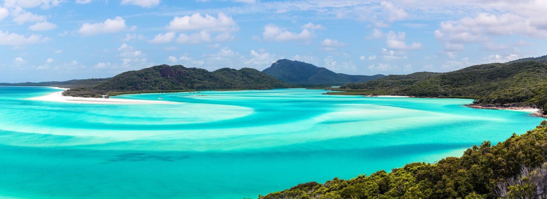 A panoramic view of a tropical beach with swirling turquoise waters and bright white sandbars, bordered by lush green forest and rolling hills. The sky above is mostly clear with scattered white clouds. A sailboat is anchored in the calm water near the shore, enhancing the serene and picturesque scene.