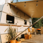 A white vintage travel trailer with brown trim parked on a hillside, featuring an extended beige awning with string lights. A wooden deck platform extends from the trailer with outdoor furniture including chairs and a small table. Potted plants and camping gear are visible on the deck. The trailer is set against a backdrop of lush green hills and mountains, with a bamboo fence visible in the background. The scene has warm, golden hour lighting suggesting an idyllic camping or glamping setup.