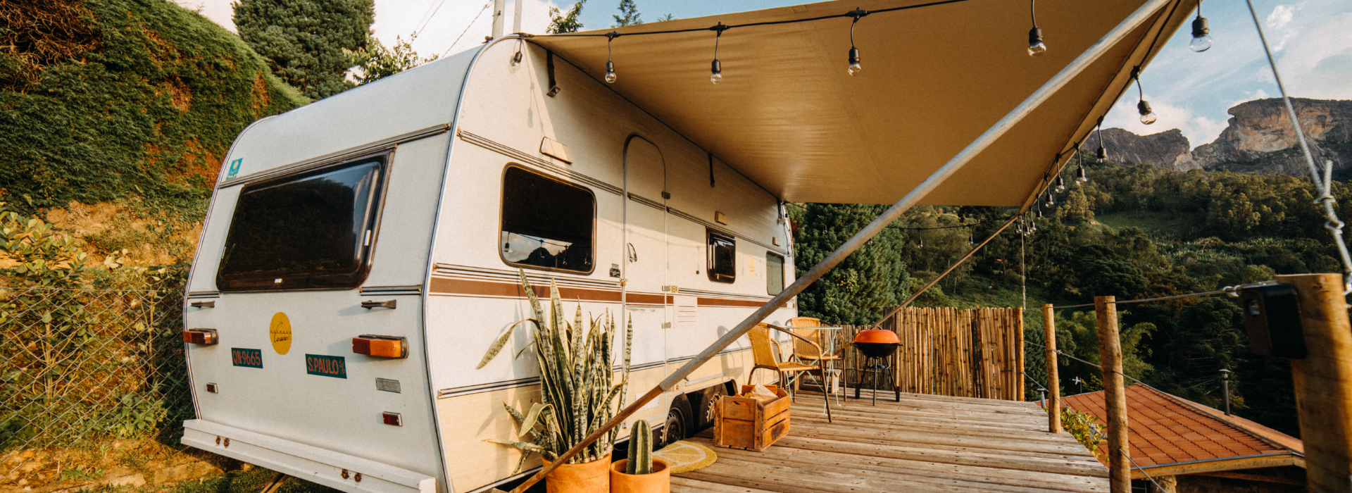 A white vintage travel trailer with brown trim parked on a hillside, featuring an extended beige awning with string lights. A wooden deck platform extends from the trailer with outdoor furniture including chairs and a small table. Potted plants and camping gear are visible on the deck. The trailer is set against a backdrop of lush green hills and mountains, with a bamboo fence visible in the background. The scene has warm, golden hour lighting suggesting an idyllic camping or glamping setup.