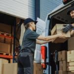 Two delivery workers unloading boxes from a commercial van outside a warehouse, representing small business operations supported by vehicle financing.
