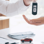A person in a white shirt handing car keys to another person across a desk, with a small red toy car, stack of documents, and pen visible on the desk surface, suggesting a car purchase or lease transaction.