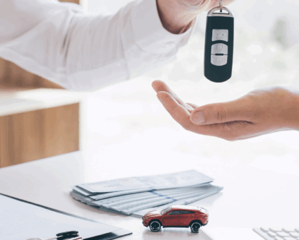 A person in a white shirt handing car keys to another person across a desk, with a small red toy car, stack of documents, and pen visible on the desk surface, suggesting a car purchase or lease transaction.