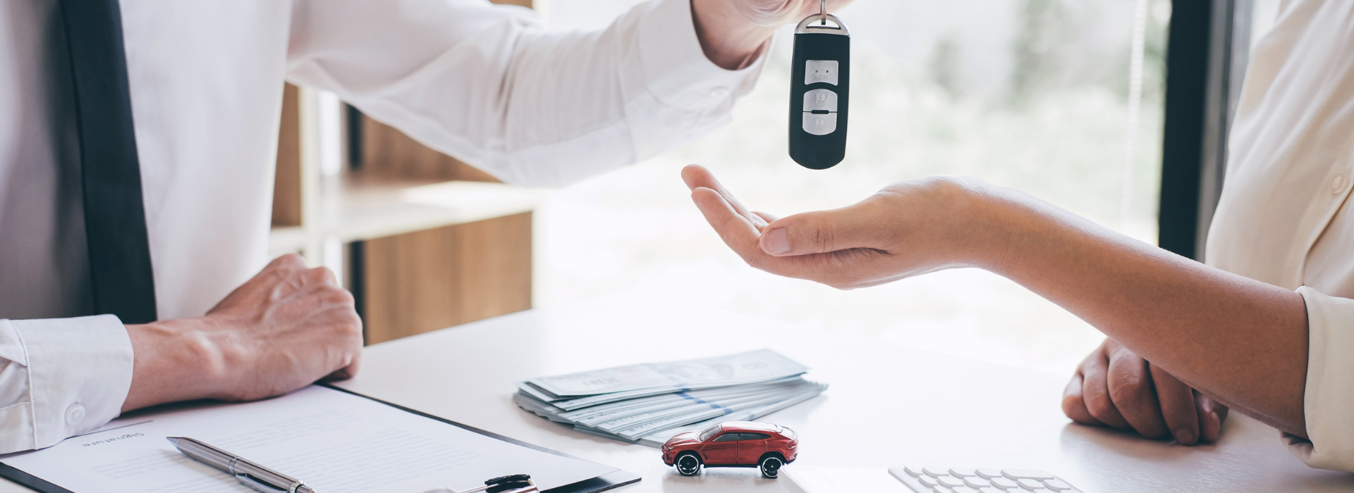 A person in a white shirt handing car keys to another person across a desk, with a small red toy car, stack of documents, and pen visible on the desk surface, suggesting a car purchase or lease transaction.