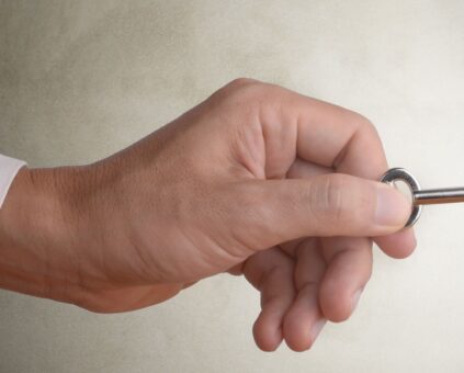 A close-up of a hand in a dark business suit sleeve with white shirt cuff holding a silver metal key. The hand is positioned as if inserting the key into a keyhole, which is visible on the right side of the image against a light-colored wall or surface. The image suggests the action of unlocking a door or gaining access