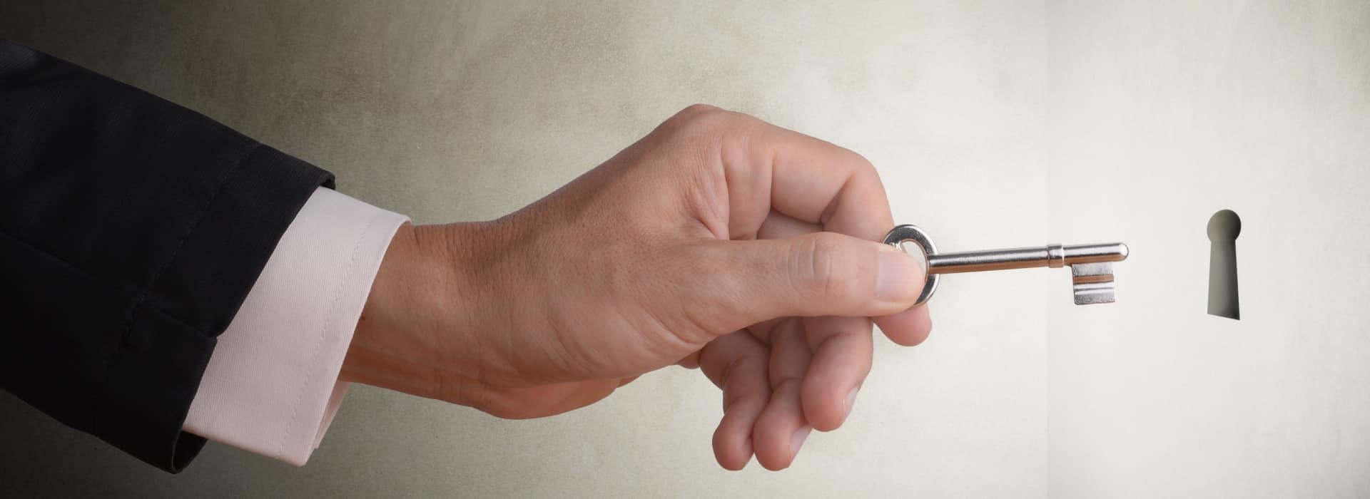 A close-up of a hand in a dark business suit sleeve with white shirt cuff holding a silver metal key. The hand is positioned as if inserting the key into a keyhole, which is visible on the right side of the image against a light-colored wall or surface. The image suggests the action of unlocking a door or gaining access