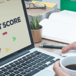 Person holding a coffee cup while using a laptop displaying 'CREDIT SCORE' text with colorful geometric shapes on the screen, sitting at a wooden desk with an open notebook, pen, and small potted plant in the background.