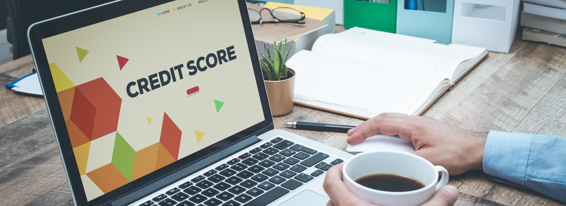 Person holding a coffee cup while using a laptop displaying 'CREDIT SCORE' text with colorful geometric shapes on the screen, sitting at a wooden desk with an open notebook, pen, and small potted plant in the background.