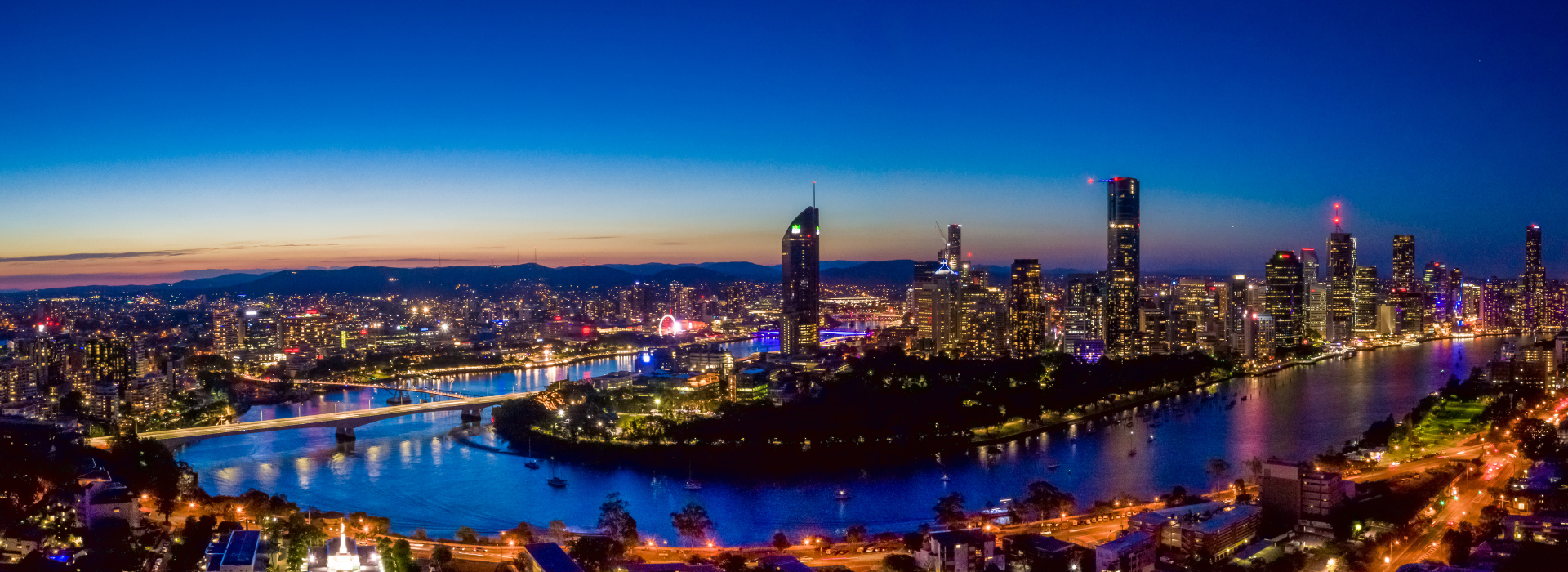 A twilight aerial view of Brisbane’s illuminated skyline, highlighting Brisbane Skytower and the winding Brisbane River, with bridges lit up and city lights reflecting off the water under a deep blue dusk sky.