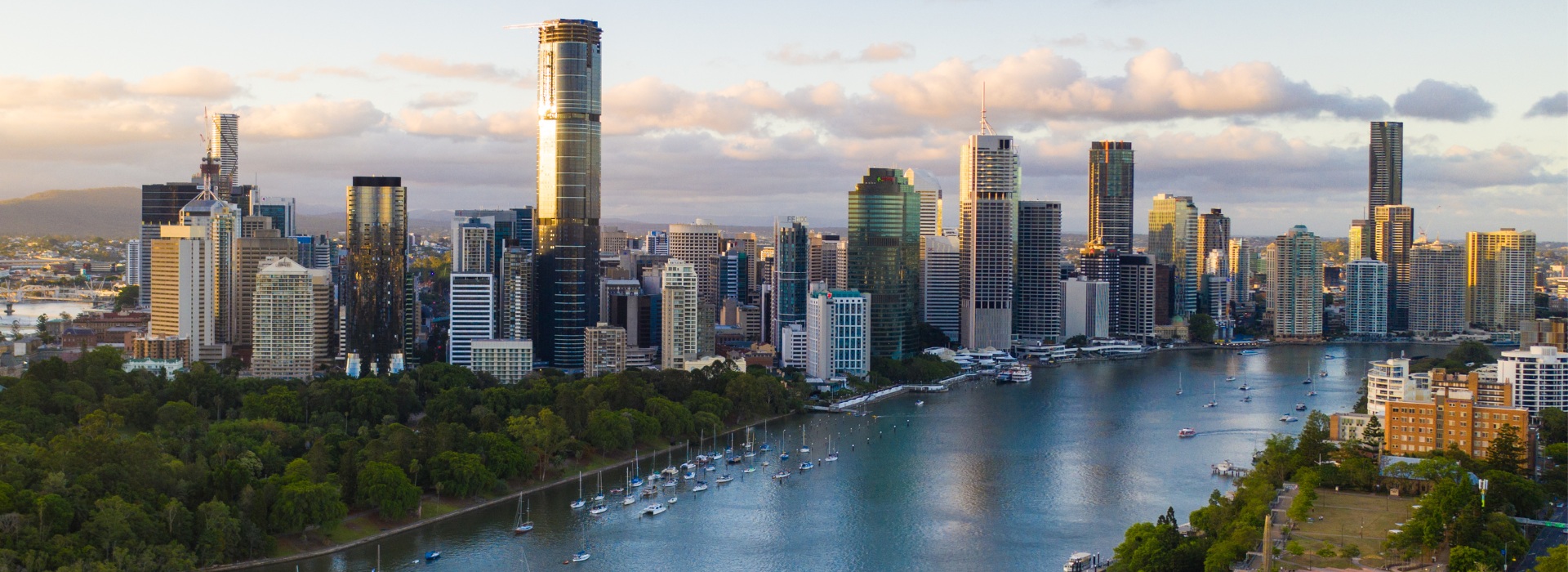 A scenic view of the Brisbane River at sunset, with a line of modern high-rise buildings along the opposite bank, and sailboats docked in the foreground.
