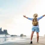 A person with a backpack and a sun hat stands on a sandy beach, with arms outstretched, facing the ocean. The image conveys a sense of freedom and adventure in a tropical setting.