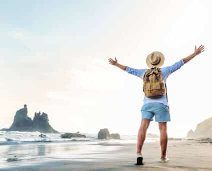 A person with a backpack and a sun hat stands on a sandy beach, with arms outstretched, facing the ocean. The image conveys a sense of freedom and adventure in a tropical setting.