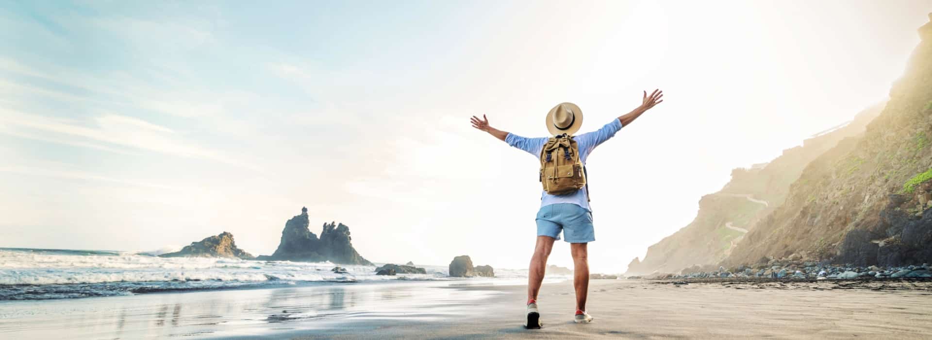 A person with a backpack and a sun hat stands on a sandy beach, with arms outstretched, facing the ocean. The image conveys a sense of freedom and adventure in a tropical setting.