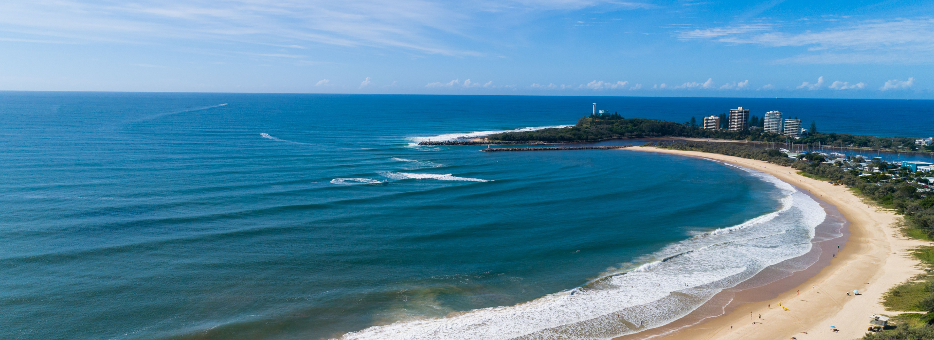  An aerial view of Mooloolaba Beach curving along the coastline, with gentle waves rolling onto the sand and a small headland with buildings and a lighthouse on the right. The vast blue ocean stretches to the horizon under a partly cloudy sky.