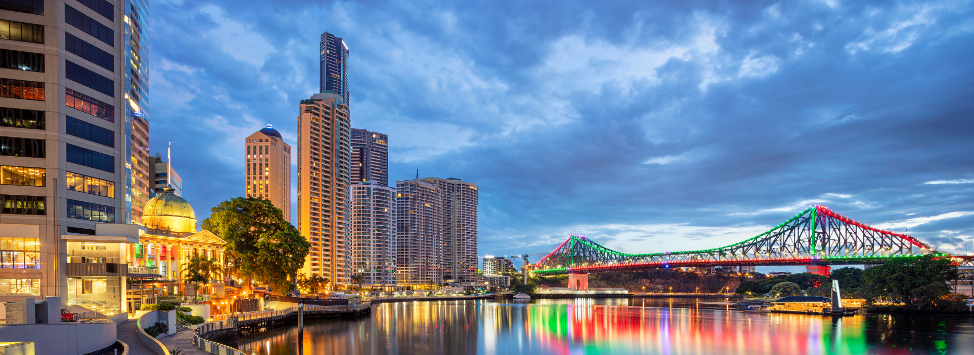  A night view of the Brisbane River with the illuminated Story Bridge and a cityscape of modern high-rise buildings and older, ornate architecture under a cloudy sky. The lights of the city and bridge are reflected in the water.