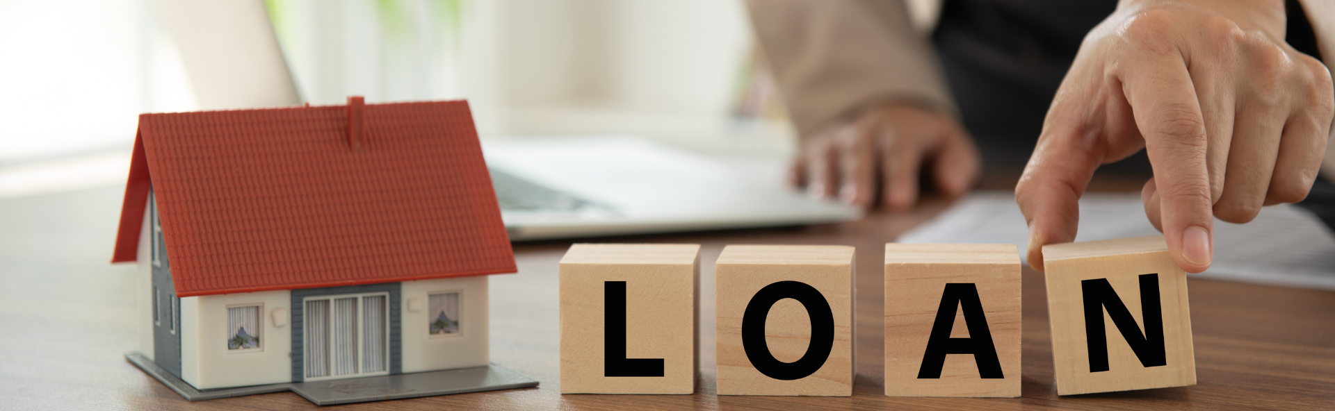 Hand arranging wooden blocks spelling 'LOAN' next to model house, representing the home loan application process