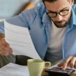 A person sitting at a kitchen table with a car loan statement and a phone, working out whether to refinance.